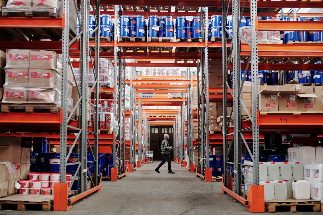 services-02 A man walking through a large industrial warehouse with stacked shelves filled with goods and products.